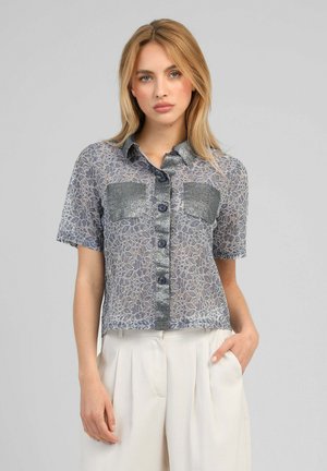 Femme portant une chemise en dentelle à manches courtes avec un motif floral et deux poches avant, associée à un pantalon blanc plissé taille haute.