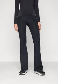 Persoon in zwarte, aansluitende flare-broek en zwart langmouwen top, staand op een effen lichtgrijze vloer en achtergrond.