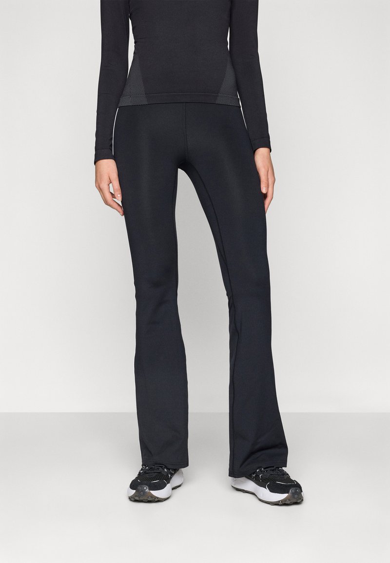 Persoon in zwarte, aansluitende flare-broek en zwart langmouwen top, staand op een effen lichtgrijze vloer en achtergrond.