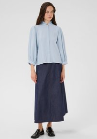 Blouse bleu clair avec un col à volants et des manches bouffantes, associée à une longue jupe en denim foncé avec une couture centrale et une coupe droite.