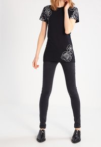 Camiseta negra con estampado floral blanco en las mangas y la parte inferior delantera. Lleva pantalones ajustados oscuros y zapatos negros.