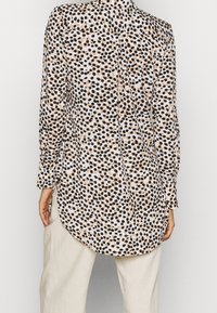 Chemisier à manches longues en tissu léger avec une base blanche et un motif à pois noir et beige. Présente un ourlet incurvé et un design avec boutons au dos.