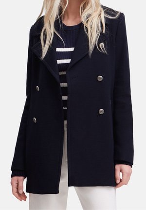 Femme portant un manteau bleu marine à double boutonnage avec des boutons argentés, par-dessus un pull rayé bleu marine et blanc et un pantalon blanc.