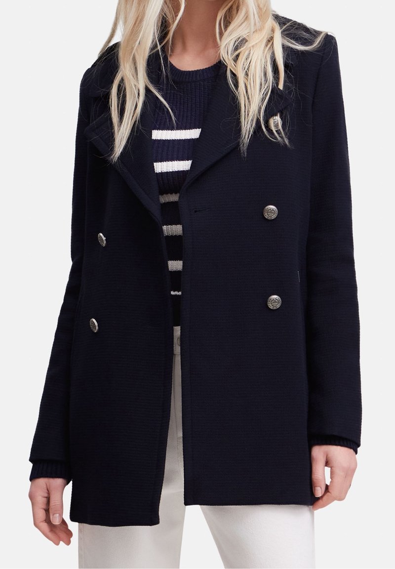 Femme portant un manteau bleu marine à double boutonnage avec des boutons argentés, par-dessus un pull rayé bleu marine et blanc et un pantalon blanc.
