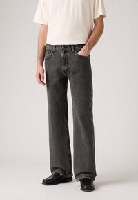 Homme portant un jean noir délavé coupe large, une chemise blanche à manches courtes, des chaussures noires et un bracelet argenté, debout avec une main dans la poche.
