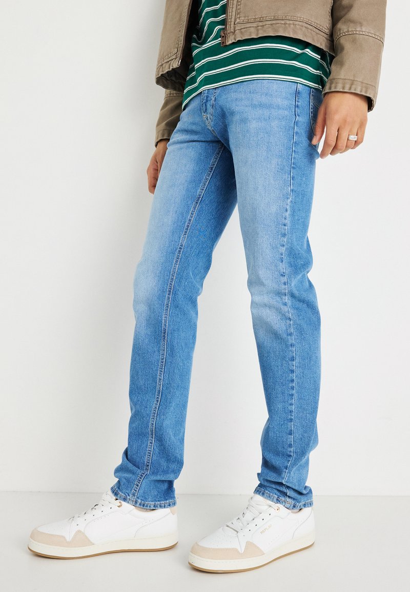 Fatos de ganga azul claro com um corte slim, apresentando uma textura ligeiramente desbotada. Combinados com sapatilhas brancas e uma camisa riscada verde.