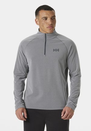 Mężczyzna o krótkich włosach i piegach, ubrany w szarą bluzę na zamek i czarne spodnie, patrzy w lewo na jednolitym tle.