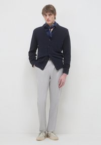 Junger Mann, der eine marineblaue Strickjacke, ein marineblaues Hemd mit Schal, hellgraue Hosen und beige Schuhe trägt, steht vor einem schlichten weißen Hintergrund.