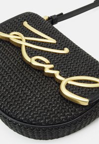 Černá tkaná kabelka kulatého tvaru s výrazným zlatým písmem loga a černým popruhem na nošení.