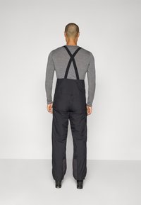 Svarta isolerade overaller med justerbara korsryggsremmar, texturerat tyg, avsmalnande ben och blixtlås vid anklarna. Grå långärmad tröja under.