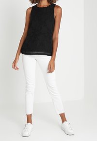 Svart ärmlös topp med texturerat blommönster, kombinerad med vita slim-fit jeans och vita sneakers, stående mot en ljus bakgrund.