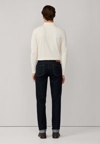 Hombre con una camisa blanca de manga larga, jeans azul oscuro con los puños remangados y zapatos de cuero marrón, de pie de espaldas sobre un fondo gris.