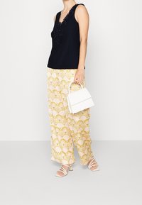 Top nero senza maniche con collo ricamato, abbinato a pantaloni larghi a fantasia nei toni crema e oro con design a ventaglio, mentre tiene una piccola borsa bianca.