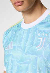 Světlomodrý sportovní dres s abstraktním vzorem, obsahuje bílý límec, tři bílé hvězdy a stříbrné logo Adidas. Hladká textura.