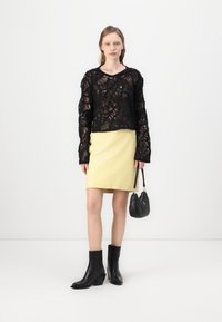 Schwarzes Spitzenoberteil mit langen Ärmeln und einem gemusterten Design, kombiniert mit einem eng anliegenden gelben Rock. Schwarze Stiefeletten und eine schwarze Handtasche vervollständigen das Outfit.
