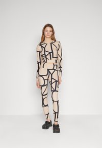 Beige treningssett med svarte geometriske mønstre. Langermet topp og matchende leggings. Modellen har på seg svarte klipklapper som fottøy.