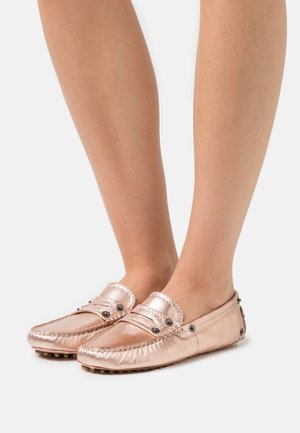 Jambes portant des mocassins en cuir métallisé rose doré avec des détails en rivets noirs, sur fond blanc.
