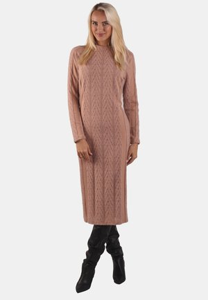 Blonde Frau in einem langen, pinkfarbenen, grob gestrickten Sweaterkleid und schwarzen, lässigen kniehohen Stiefeln, die mit überkreuzten Beinen steht.