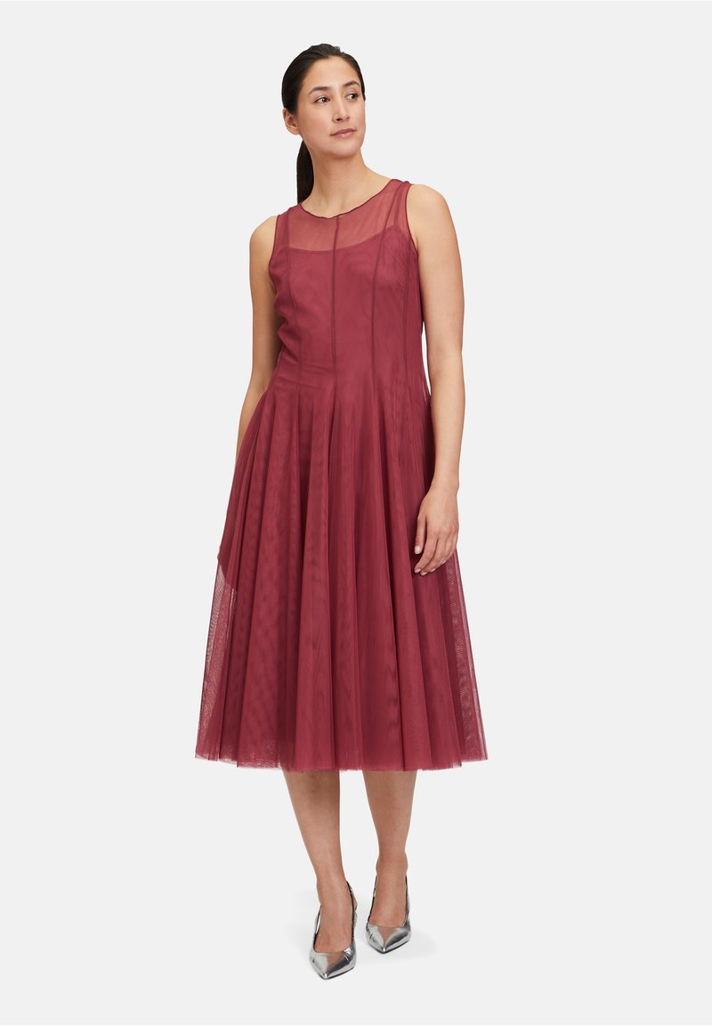 Bordeauxfarbenes Kleid mit tailliertem Oberteil und ausgestellt Rock, aus leichtem Material mit transparenter Überlagerung. Silberne spitze Absätze runden das Outfit ab.