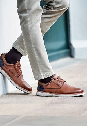 Osoba nosząca beżowe spodnie i brązowe skórzane sznurowane buty z czarnymi skarpetkami, stojąca z jedną stopą opartą na krawędzi na zewnątrz.