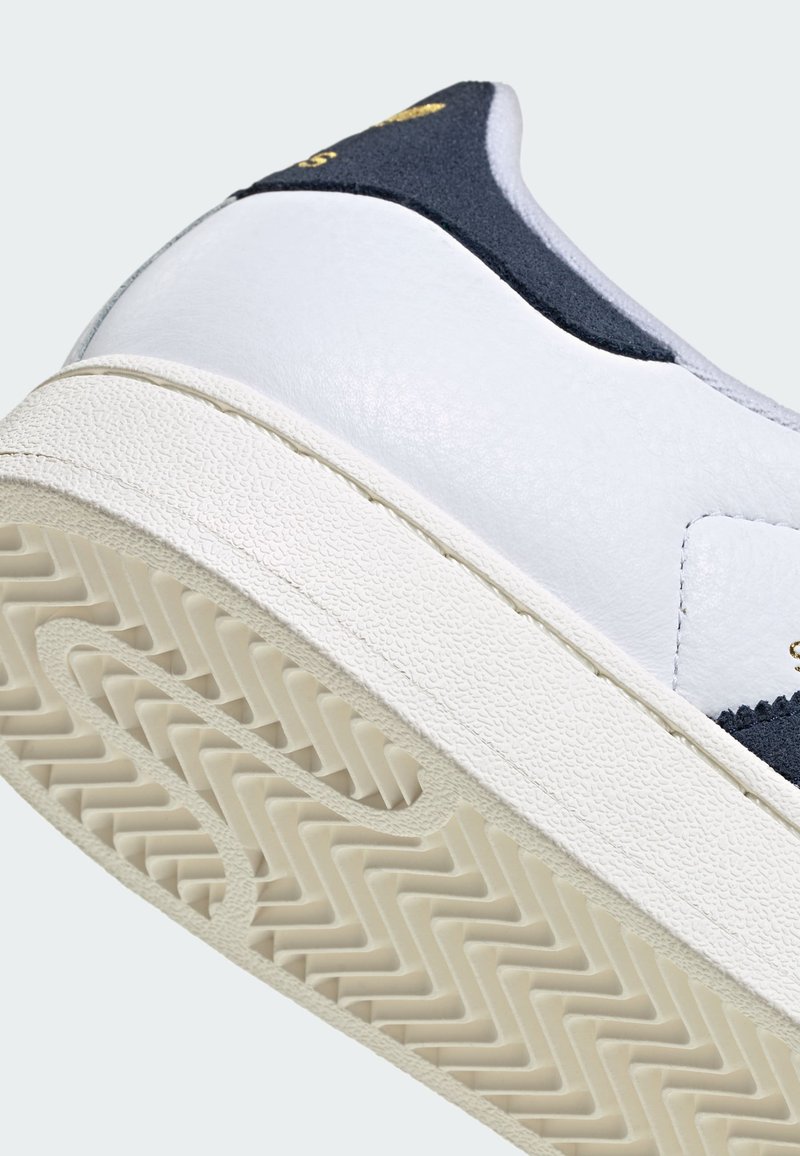White sneaker with textured rubber sole, navy blue heel accent, and gold branding on heel tab.