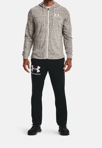 Grå zip-up hoodie med snoretræk, båret over sorte bukser med et hvidt logo; parret med sorte træningssko. Glat tekstur.