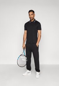 Černé polo tričko s bílým límečkem, spárované s texturovanými černými tepláky. Model drží tenisovou raketu a má na sobě bílé sportovní boty.