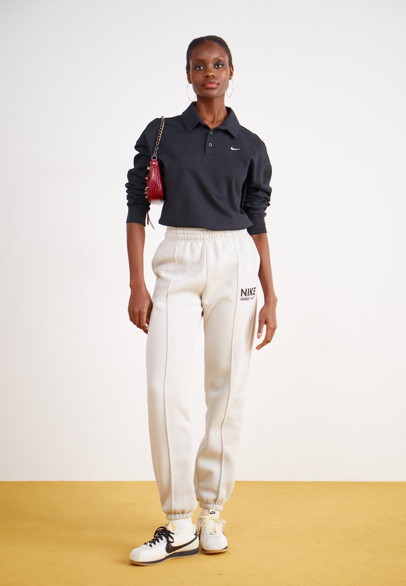 Camisa polo negra de manga larga, pantalones de chándal blancos con el logo de Nike y zapatillas blancas. El accesorio incluye un pequeño bolso rojo.
