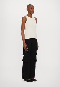 Top sin mangas de canalé crema combinado con pantalones cargo negros que cuentan con múltiples bolsillos, diseño de pierna recta y zapatos planos negros.