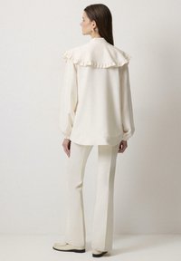 Blusa crema con cuello con volantes, mangas largas y puños elásticos, combinada con pantalones acampanados a juego y zapatos ligeros. Textura suave.