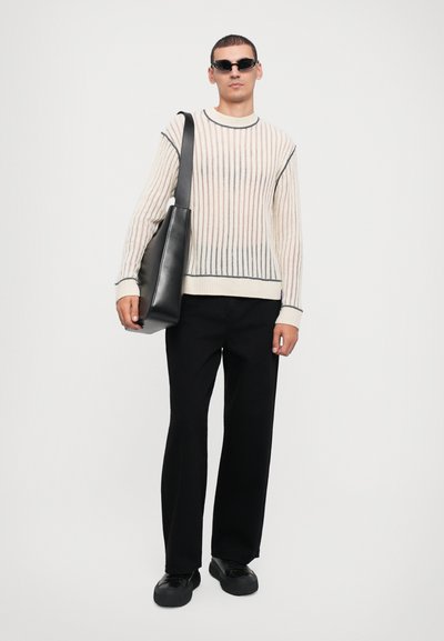 Knit sweater with vertical stripes in cream and gray, paired with black wide-leg trousers and a black tote bag. Model wears dark sunglasses.