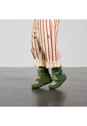 Kind trägt cremefarbene Hose mit roten Streifen und grüne High-Top-Schuhe, steht auf einem glatten grauen Boden vor einem einfarbigen Hintergrund.