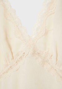 Kremasto obojena tkanina s valovitim cvjetnim čipkastim obrubom koji oblikuje V-izrez i presijecajući detalj na laganom odjevnom predmetu.