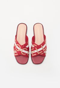 Les sandales rouges à semelle plate présentent un design à brides croisées avec une texture tissée, agrémentées d'un motif de logo beige et d'une semelle intérieure lisse.