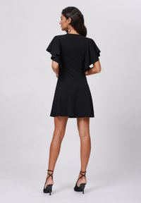 Robe noire courte avec des manches flottantes, un corsage ajusté et une jupe évasée. Portée avec des talons hauts à lanières noires. Texture de tissu lisse.