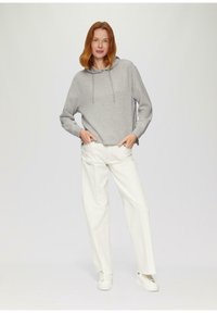 Felpa grigia con cappuccio e stringhe, abbinata a pantaloni bianchi a gamba larga. Il modello sta in piedi con le mani nelle tasche, indossando sneakers bianche.