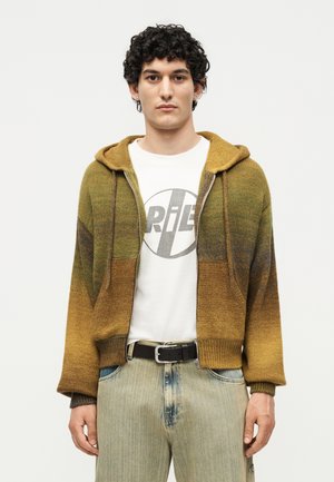 Ein junger Mann mit lockigem Haar trägt einen braun-grünen Ombré-Zip-Hoodie über einem weißen T-Shirt mit Grafik und ausgewaschenen blauen Jeans.