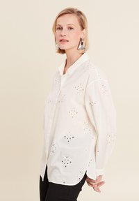 White button-up shirt with embroidered cutouts in floral patterns. Long sleeves and curved hem; made of lightweight fabric.