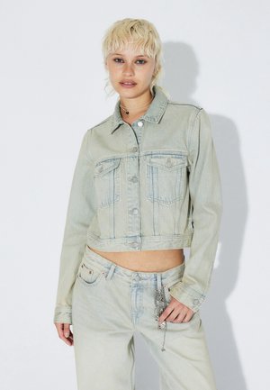 Veste courte en denim bleu clair avec manches longues, fermeture à boutons, deux poches poitrine et coutures contrastantes. Portée avec un pantalon taille haute assorti.