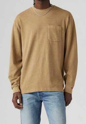 Homme portant une chemise Levi’s beige à manches longues avec poche poitrine et un jean bleu clair, debout devant un fond clair uni.