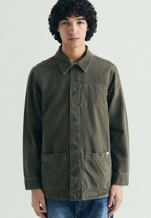 Joven con cabello rizado, vestido con una chaqueta verde oliva abotonada con tres bolsillos delanteros y jeans azules, de pie contra un fondo liso.