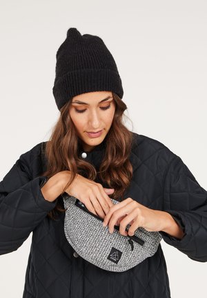 Gequilted zwarte jas, gebreide zwarte beanie en een ronde grijze tas met textuur, met een zwarte rits en logo, die door een persoon wordt geopend.