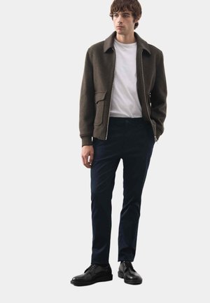 Veste olive à fermeture éclair avec un col, portée avec un t-shirt blanc, un pantalon bleu marine et des chaussures noires. Design minimaliste avec une coupe structurée.