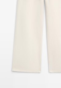 Parte inferiore di pantaloni color crema a gamba dritta con cuciture laterali visibili e orli rifiniti su sfondo bianco.