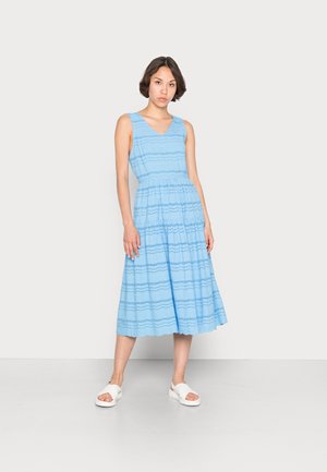 Vaaleansininen hihaton mekko, jossa on V-pääntie, vaakasuorat kuviot ja liehuvahelma. Yhdistetty valkoisiin sandaaleihin.