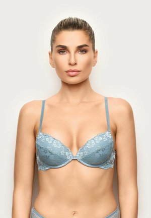 Donna con capelli legati, che indossa un reggiseno di pizzo azzurro chiaro, rivolta in avanti su uno sfondo neutro e uniforme.