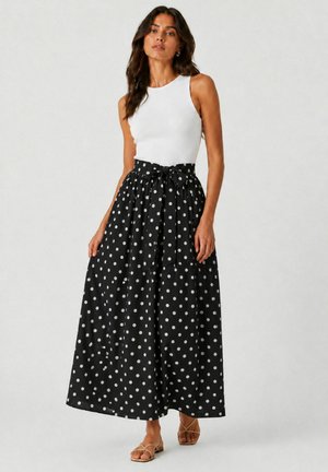 Donna che indossa una canotta bianca senza maniche e una gonna lunga nera a vita alta con pois bianchi e cintura annodata, con sandali beige.