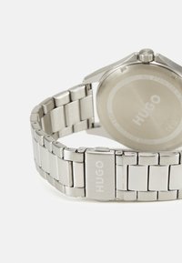 Reloj de acero inoxidable plateado con pulsera de eslabones, que presenta una caja redonda y un logo grabado en el cierre y en la parte trasera.
