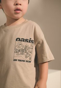 T-shirt en coton beige avec un imprimé graphique noir représentant une scène avec des personnes, des arbres et des véhicules. Le texte dit "oasis SOIS ICI MAINTENANT."