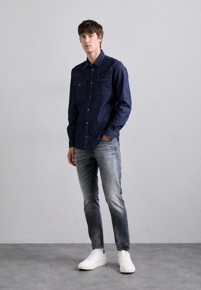 Camicia in denim scuro con bottoni e due taschini sul petto, abbinata a jeans grigi sbiaditi e sneakers bianche, su uno sfondo neutro.